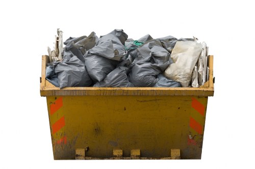 A large yellow metal skip filled with tightly packed black and grey rubbish bags, some white plastic items, and assorted debris, situated outdoors on a concrete surface. The skip displays orange reflective safety stripes on its sides and has signs of dirt and wear. The environment appears to be a waste collection area, potentially near a commercial or residential property in Pimlico, with no visible background details. The image captures the typical scene associated with rubbish removal services, such as those provided by Office Clearance Pimlico, highlighting the process of discarding mixed waste materials in a container ready for collection.