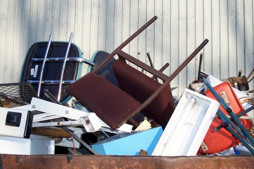A collection of discarded items piled together outdoors in front of a corrugated metal wall, including a dark blue plastic chair, a brown upholstered armchair with wooden legs, a white plastic frame or panel, a blue storage bin, and various metal and plastic components. The items are stacked haphazardly on a rusted metal surface or platform, with some objects leaning or overlapping others. The scene appears to be part of a rubbish removal service operation, typical of waste collected for disposal or recycling in an urban environment near Pimlico. The natural lighting highlights the textures and colors of the different materials, emphasizing the mixture of plastic, wood, and metal waste, which are common in office clearance and rubbish removal contexts handled by Office Clearance Pimlico.