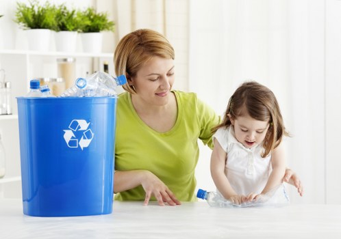 A woman with short, light brown hair, wearing a bright yellow-green top, is sitting at a table with a young girl who has long, light brown hair tied back and is dressed in a white short-sleeved shirt. They are both engaged in a waste disposal activity, with the woman guiding the girl who is handling a clear plastic bottle. To the left of the woman is a large blue recycling bin made of plastic with a white recycling symbol on its surface, filled with crushed plastic bottles and other plastic waste. The background features a white shelving unit with potted green plants, creating a bright and tidy indoor environment. The scene emphasizes responsible rubbish management, subtly aligning with the services of Office Clearance Pimlico in the local area, highlighting environmentally conscious disposal practices within a domestic or office setting.