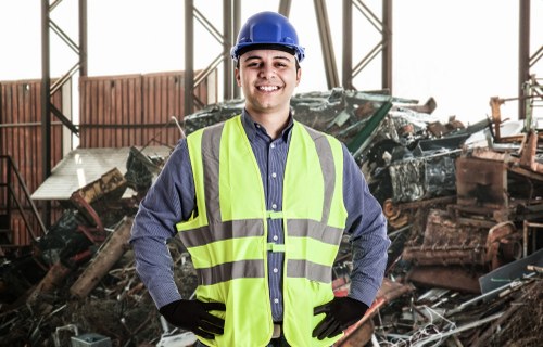 A young male worker wearing a blue safety helmet, a high-visibility yellow vest, and black gloves stands inside an industrial warehouse with metal framework. Behind him is a pile of assorted rubbish, including broken wooden furniture, metal scrap, and other discarded materials, indicating a rubbish removal or clearance scene. The environment is well-lit, with natural light filtering through the large windows or open structural elements. The worker is smiling confidently, conveying professionalism and readiness to handle waste management tasks. This image emphasizes the type of rubbish removal services offered by Office Clearance Pimlico in central London, particularly in areas like Pimlico and surrounding postcode districts, capturing a professional approach to clearing waste from commercial or industrial sites.