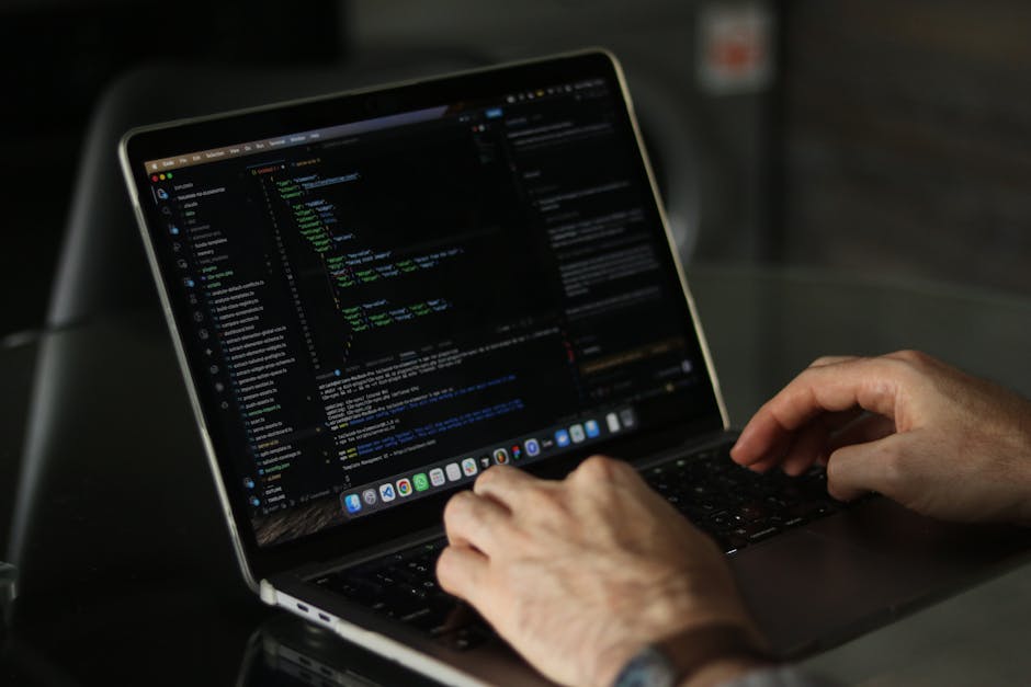 A person with dark hair, dressed in a light blue shirt, is sitting at a desk in an office environment, working on a silver laptop with a black keyboard. The laptop screen displays colorful lines of co