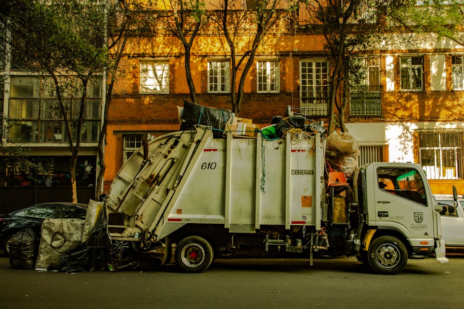 A commercial waste collection truck parked on a city street with its rear hydraulic lift open, revealing a large, open-top steel compartment filled with various types of rubbish, including black plast