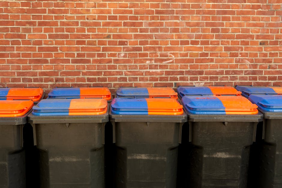 Two large black plastic rubbish bags, visibly crumpled and tied at the top, are placed directly on a paved sidewalk next to a black metal fence. The bags appear to be filled with waste materials, with