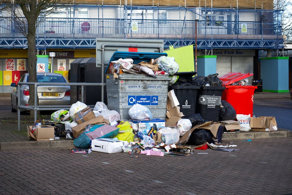 A collection of multiple plastic rubbish bags, primarily in yellow, white, and transparent colours, are piled on a cobblestone pavement against a white-painted brick wall with curved architectural det