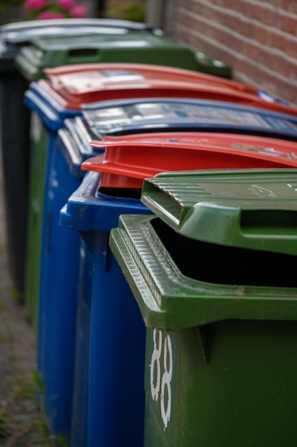 A row of large plastic wheelie bins placed along a pavement beside a brick wall. The bins are arranged in a sequence, with green bins at the front, followed by blue, red, and dark green bins further d