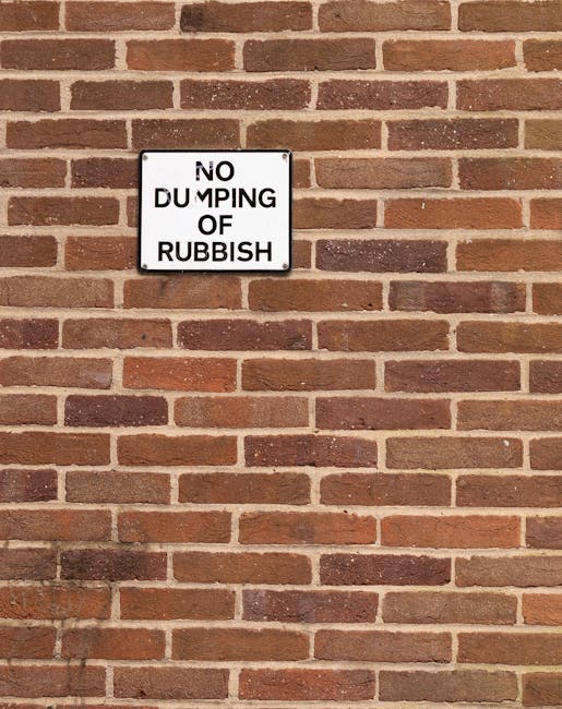 A row of large plastic wheelie bins placed along a pavement beside a brick wall. The bins are arranged in a sequence, with green bins at the front, followed by blue, red, and dark green bins further d