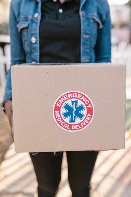A person dressed in a dark t-shirt and a blue denim jacket is standing outdoors, holding a cardboard box with both hands. The box features a prominent circular sticker displaying a blue medical symbol