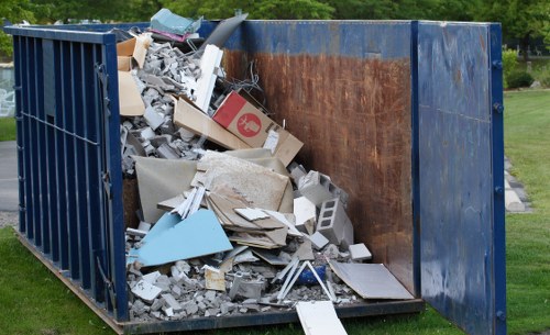 A large open-top skip situated outdoors on a grassy area, filled with a mixture of construction and household waste. The contents include broken drywall, discarded wooden panels, a cardboard box with a red logo, broken tiles, small pieces of plaster, and various other debris. The skip's sides are made of dark blue metal, with one side showing rust and weathering, indicating exposure to the elements. The scene appears to be part of a waste removal or clearance operation in an urban or suburban environment, consistent with services offered by Office Clearance Pimlico. The background includes a well-maintained lawn and some trees, suggesting the skip is placed on a residential or park-like site in Pimlico or nearby in Westminster.