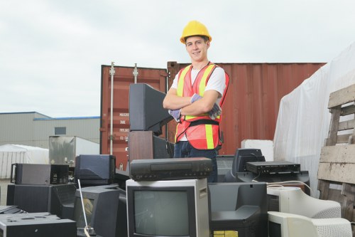A man wearing a yellow safety helmet and a high-visibility vest stands outdoors amidst a collection of discarded electronic appliances, including old computer monitors, keyboards, and other obsolete electronic devices, which are stacked and scattered on the ground. The background features a shipping container and industrial warehouse structures under a cloudy sky. The scene appears to take place on a paved or gravel surface, likely in an area designated for waste or electronic disposal, illustrating electronic rubbish removal. The individual shows a confident posture with arms crossed, and the overall environment suggests a waste management or recycling operation, consistent with rubbish removal services provided by companies like Office Clearance Pimlico serving the Pimlico area.