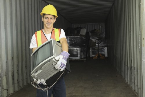 A young man wearing a yellow safety helmet, a reflective high-visibility vest, white shirt, and work gloves, standing inside a storage container with metal walls. He is holding an old, bulky computer monitor with a grey casing and a curved screen, positioned at waist height. The container appears to be used for storage or disposal, with stacked electronic waste visible in the background. The scene is outdoors, with natural light illuminating the interior, related to rubbish removal services in Pimlico. The image emphasizes environmentally responsible disposal practices and aligns with professional office clearance services offered by Office Clearance Pimlico in Westminster, London, with careful handling of electronic waste.