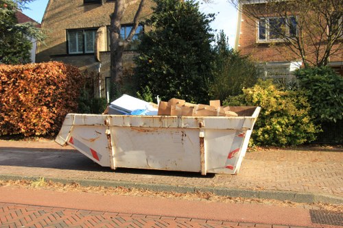 A large, white, metal skip filled with various cardboard boxes and packaging materials, situated on a residential street pavement in Pimlico. The skip shows signs of rust and wear, with some edges slightly bent or corroded. It is positioned close to the curb, adjacent to a brick-paved driveway, with neatly trimmed bushes and trees in the background, along with residential houses featuring brick and stone exteriors and multiple windows. The scene is illuminated by daylight, indicating a clear, sunny day. This image relates to rubbish removal services offered by Office Clearance Pimlico, emphasizing collection and disposal of waste from residential properties in the Pimlico area.