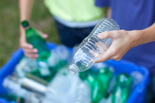 A close-up of a person's hand holding a clear plastic bottle over a blue recycling bin filled with various plastic bottles and containers, some green and others transparent, with visible ridges and caps. In the background, two other individuals are partially visible, one with a green shirt and the other with a blue top, near a paved or concrete surface likely outdoors. The scene depicts a waste recycling activity, emphasizing plastic waste collection, relevant to rubbish removal services in Pimlico. The environment appears well-lit, and the focus is on the plastic bottle being held, illustrating the process of sorting recyclable materials as part of office clearance or general rubbish disposal efforts.
